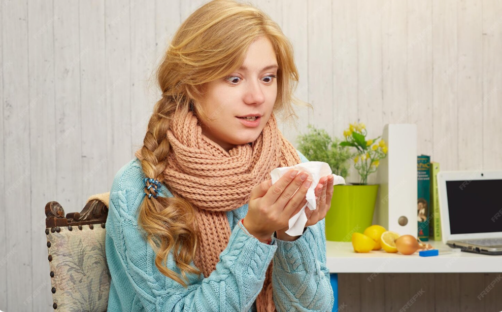 Woman with cold sore holding tissue, wearing a scarf, near laptop and plants